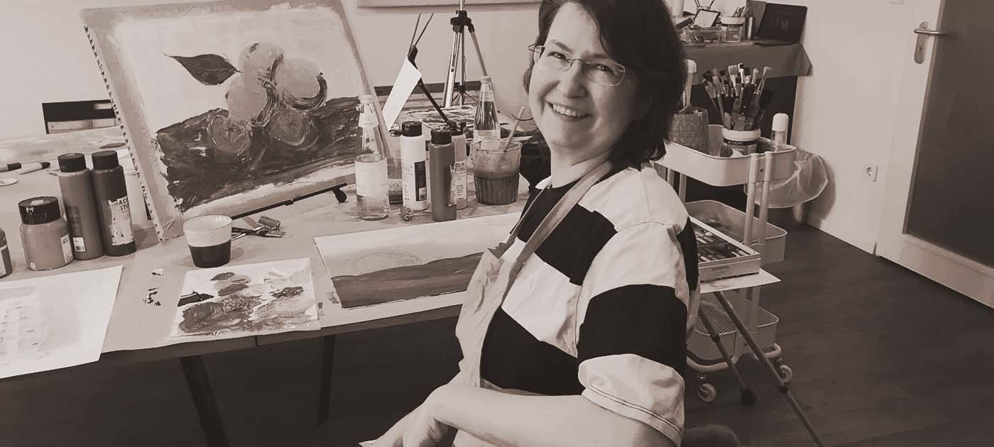 Ein Sepiabild. Eine dunkelhaarige Frau mit Brille schaut den Betrachter direkt an. Sie sitzt vor einer Staffelei mit einem Früchtestillleben in einem Atelier.